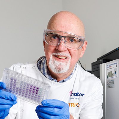 a man in a white coat, glue gloves and glasses holds up a small tray of purple dots